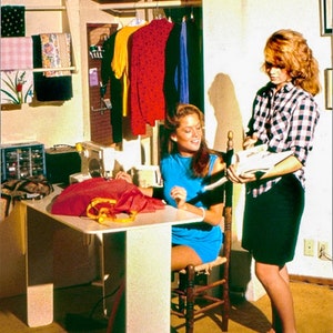 May include: Two people are working on a sewing project. One person is sitting at a white table with a sewing machine and fabric. The other person is standing and holding a piece of fabric. The room is decorated with a wooden closet and shelves with fabric and clothing.