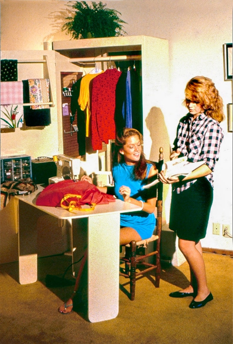 May include: Two people are working on a sewing project. One person is sitting at a white table with a sewing machine and fabric. The other person is standing and holding a piece of fabric. The room is decorated with a wooden closet and shelves with fabric and clothing.