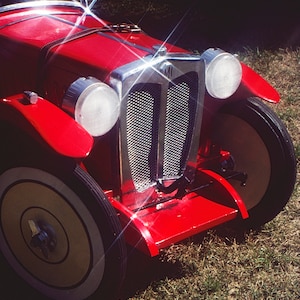 Puede incluir: Un coche de juguete rojo con una parrilla cromada y faros. El coche tiene un volante blanco y un asiento de cuero negro. El coche est&aacute; sentado en un parche de hierba verde.