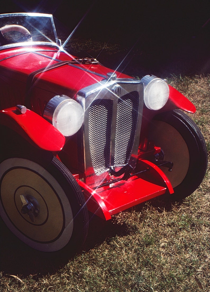 Puede incluir: Un coche de juguete rojo con una parrilla cromada y faros. El coche tiene un volante blanco y un asiento de cuero negro. El coche est&aacute; sentado en un parche de hierba verde.