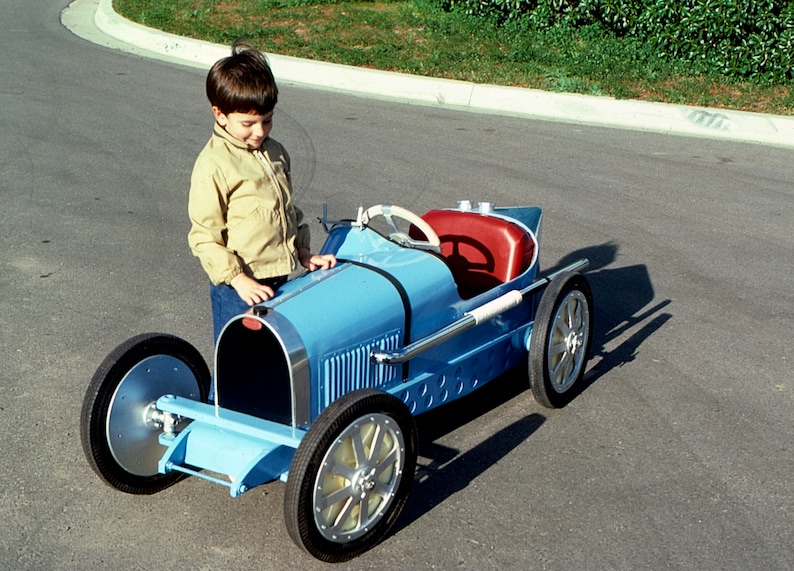 May include: A young boy stands next to a blue toy car that resembles a classic Bugatti. The car has a red leather seat and a steering wheel. The boy is wearing a tan jacket and blue jeans.