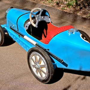 May include: A blue toy car shaped like a vintage race car. The car has a steering wheel, a red seat, and black tires.