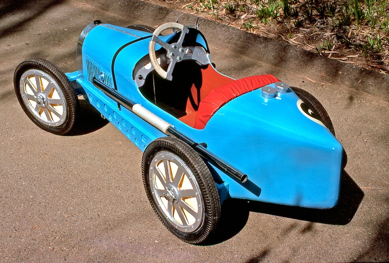May include: A blue toy car shaped like a vintage race car. The car has a steering wheel, a red seat, and black tires.
