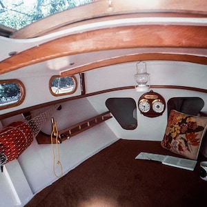 May include: The interior of a small boat with white walls and wooden trim. There is a red and white net hanging from the ceiling, a cushion with a floral pattern on the bench, and a clock on the wall.