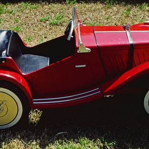 Puede incluir: Un coche de juguete rojo con un asiento de cuero negro y ruedas con llantas blancas. El coche tiene un parachoques cromado y una correa de cuero negro sobre el cap&oacute;.