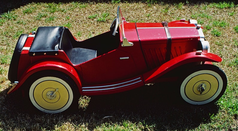 Puede incluir: Un coche de juguete rojo con un asiento de cuero negro y ruedas con llantas blancas. El coche tiene un parachoques cromado y una correa de cuero negro sobre el cap&oacute;.