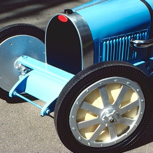 May include: A blue toy car with a black grill and silver accents. The car has a large, silver steering wheel and black tires.