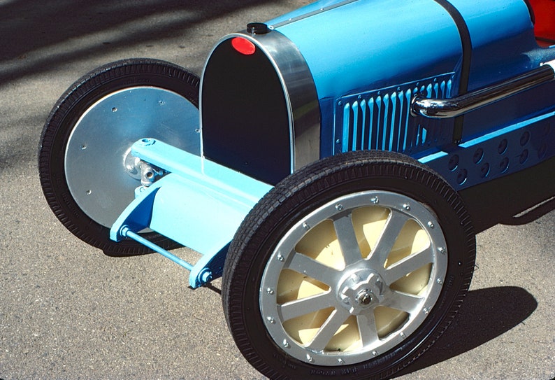 May include: A blue toy car with a black grill and silver accents. The car has a large, silver steering wheel and black tires.