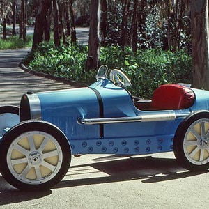 May include: A blue toy car shaped like a vintage Bugatti race car. The car has a red seat and a steering wheel. The car is on a paved road with trees in the background.