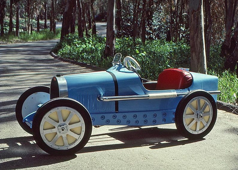 May include: A blue toy car shaped like a vintage Bugatti race car. The car has a red seat and a steering wheel. The car is on a paved road with trees in the background.