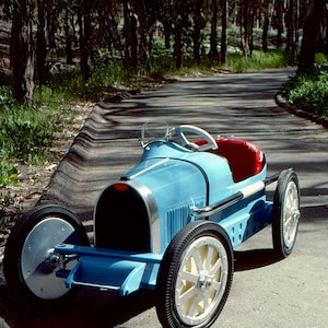 May include: A blue toy car shaped like a vintage race car. The car has a red seat, a steering wheel, and four black wheels with white spokes. The car is on a paved road with trees in the background.