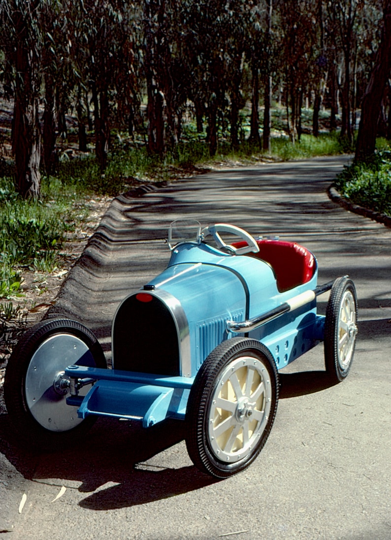 May include: A blue toy car shaped like a vintage race car. The car has a red seat, a steering wheel, and four black wheels with white spokes. The car is on a paved road with trees in the background.