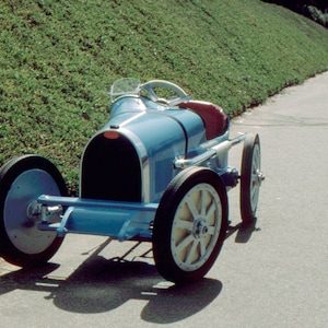 May include: A blue toy car with a black grill and white wheels. The car is on a paved road with a grassy hill in the background.