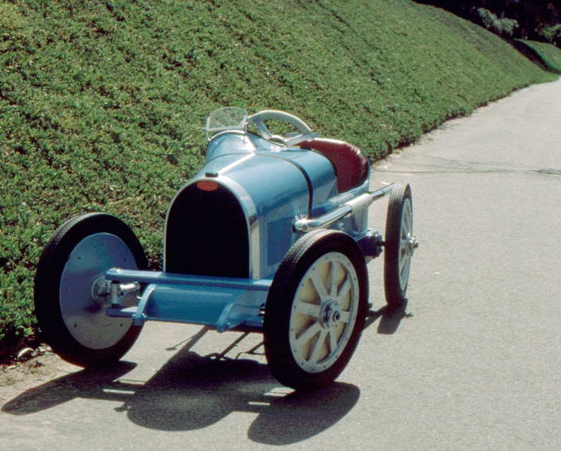 May include: A blue toy car with a black grill and white wheels. The car is on a paved road with a grassy hill in the background.