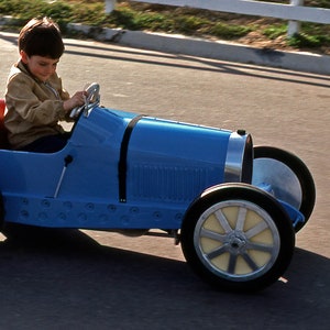 May include: A young child drives a blue toy car with a number "2" on the side. The car has a black steering wheel and white wheels.