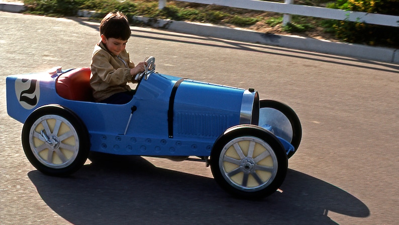 May include: A young child drives a blue toy car with a number "2" on the side. The car has a black steering wheel and white wheels.