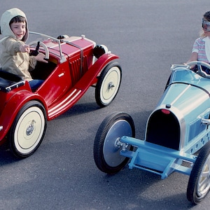 May include: Two children are driving toy cars. The boy is driving a red toy car with a black spare tire on the back. The girl is driving a blue toy car with a black grill. Both cars have white tires.