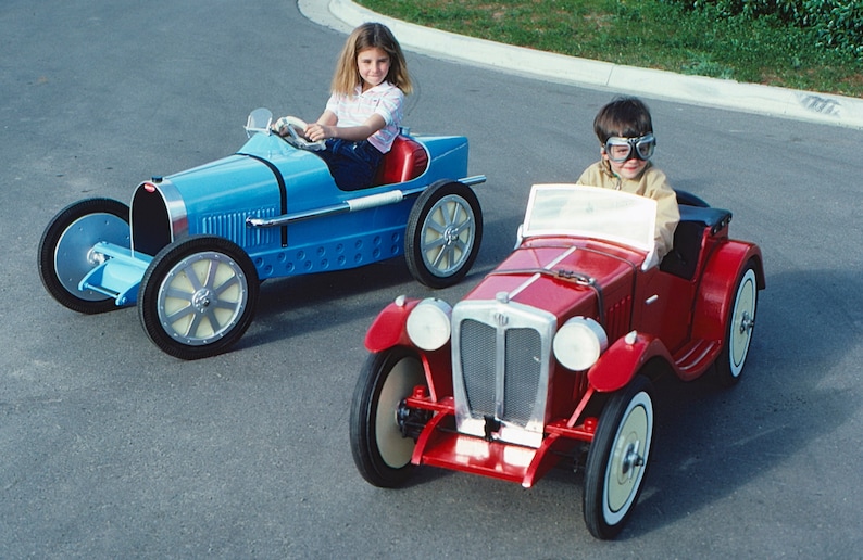 May include: Two children are driving toy cars. The girl is driving a blue toy car with a white steering wheel. The boy is driving a red toy car with a white steering wheel. Both cars have white tires.