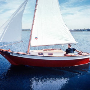 May include: A red and white sailboat with a white sail is on the water. The boat has a wooden mast and a wooden bowsprit. The boat is sailing with the wind. The boat has a registration number "CA 4287 FL" on the side.