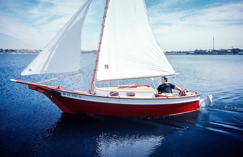 May include: A red and white sailboat with a white sail is on the water. The boat has a wooden mast and a wooden bowsprit. The boat is sailing with the wind. The boat has a registration number "CA 4287 FL" on the side.