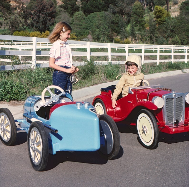 Puede incluir: Dos ni&ntilde;os jugando con coches de juguete. Una ni&ntilde;a con una camisa a rayas rosa y blanca est&aacute; de pie junto a un coche de juguete azul. Un ni&ntilde;o con una chaqueta marr&oacute;n est&aacute; sentado en un coche de juguete rojo.