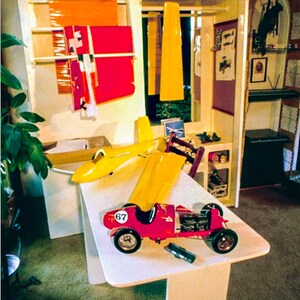 May include: A red and yellow toy race car with the number 67 on the side, a yellow toy airplane, and a yellow and red toy plane on a white table. The toys are in a room with a wooden shelf unit in the background.