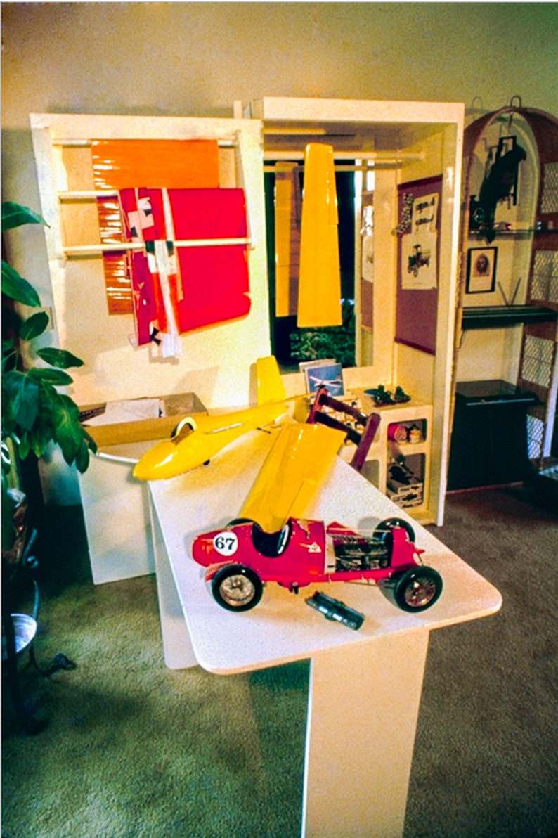 May include: A red and yellow toy race car with the number 67 on the side, a yellow toy airplane, and a yellow and red toy plane on a white table. The toys are in a room with a wooden shelf unit in the background.