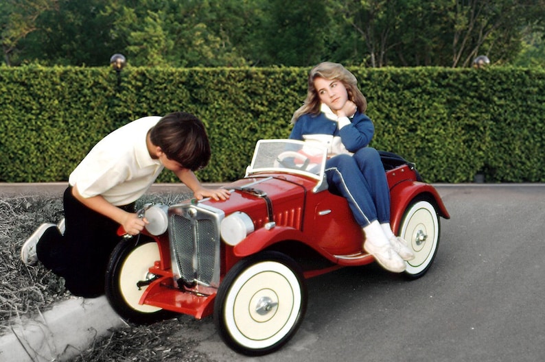 Puede incluir: Un coche de juguete rojo con un volante blanco y neum&aacute;ticos blancos. Un ni&ntilde;o est&aacute; arrodillado junto al coche y una ni&ntilde;a est&aacute; sentada en el asiento del conductor.