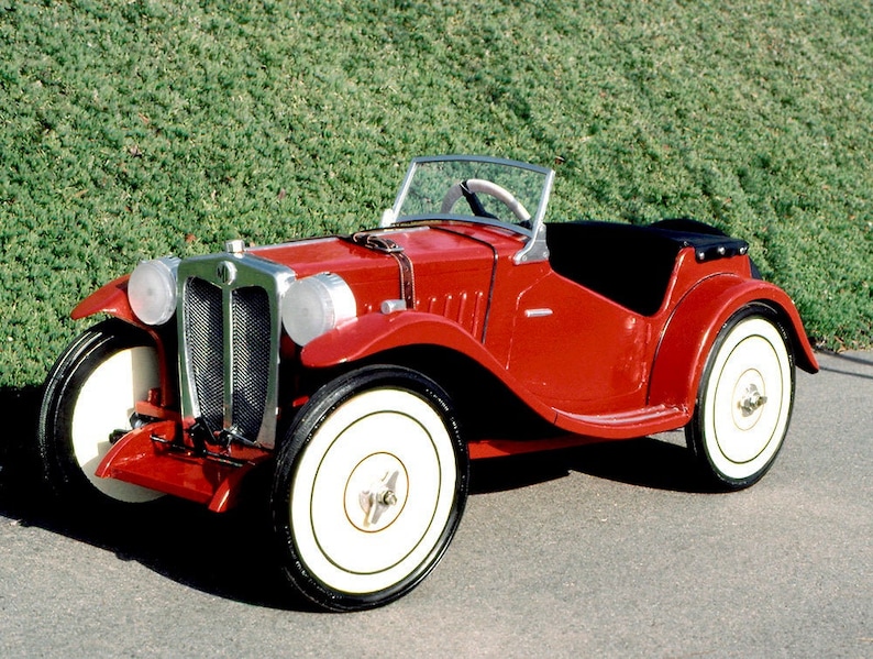 Puede incluir: Un coche de juguete vintage rojo con neum&aacute;ticos de banda blanca y capota de tela negra. El coche tiene una parrilla y faros cromados.