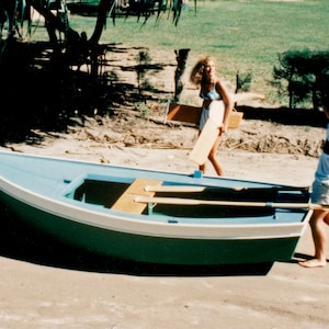 May include: Two people are carrying a green and white wooden rowboat on a sandy beach. The boat has two wooden oars inside.