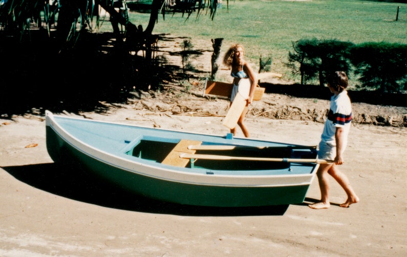 May include: Two people are carrying a green and white wooden rowboat on a sandy beach. The boat has two wooden oars inside.