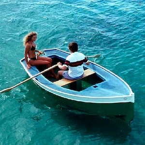 May include: Two people in a small blue and white rowboat on a clear blue body of water. The boat is being propelled by a wooden oar.