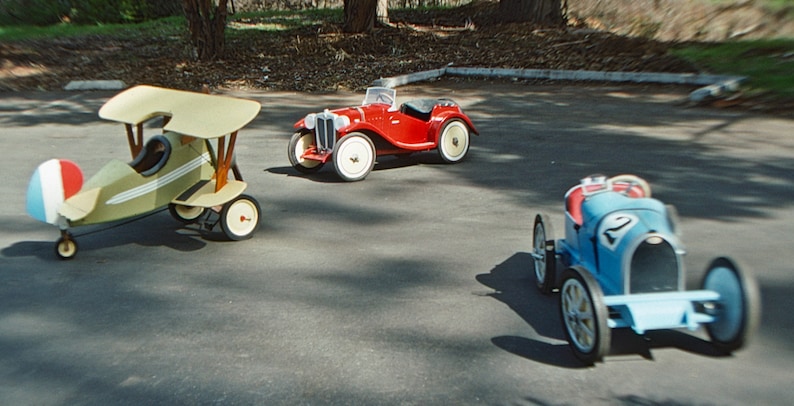 Puede incluir: Tres veh&iacute;culos de juguete, un roadster rojo, un coche de carreras azul y un biplano verde y blanco, est&aacute;n alineados en una superficie de asfalto gris.