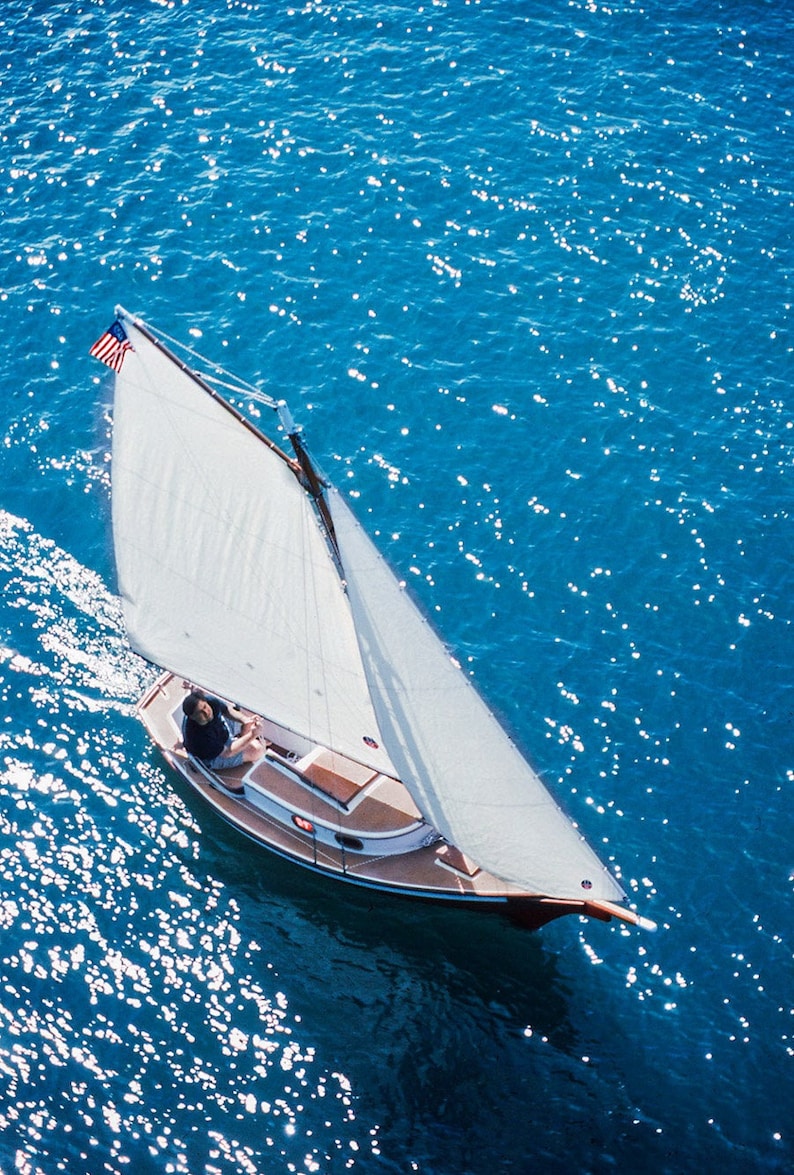 May include: A small sailboat with a white sail and a wooden hull is sailing on a blue body of water. The sailboat has an American flag on the mast. A person is sitting in the cockpit of the boat.
