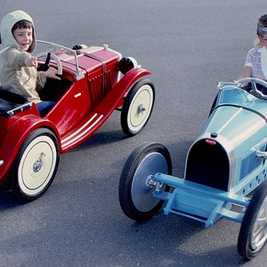 Puede incluir: Dos ni&ntilde;os con atuendos de carreras vintage conducen coches de juguete. El ni&ntilde;o con un casco blanco conduce un coche rojo, mientras que la ni&ntilde;a con gafas de protecci&oacute;n conduce un coche azul.