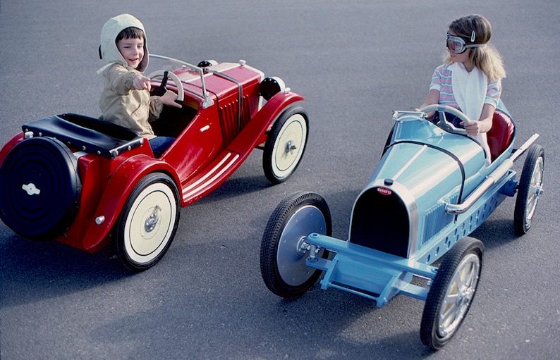 Puede incluir: Dos ni&ntilde;os con atuendos de carreras vintage conducen coches de juguete. El ni&ntilde;o con un casco blanco conduce un coche rojo, mientras que la ni&ntilde;a con gafas de protecci&oacute;n conduce un coche azul.