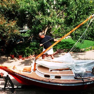 May include: A small wooden sailboat with a white sail, sitting on a trailer. The boat is red and white with a wooden mast and a wooden rudder. A person is standing near the boat, holding a rope.