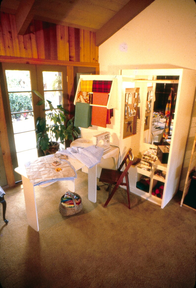 May include: A sewing room with a white sewing machine, a wooden chair, and a white cabinet with shelves. The cabinet is filled with fabric scraps, spools of thread, and other sewing supplies. There is a table with a blue and white checkered tablecloth and a basket of fabric scraps.