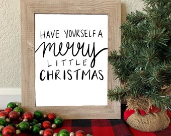 Peut inclure: Une impression encadrée avec le texte "Have yourself a merry little Christmas" en écriture noire sur fond blanc. L'impression est posée sur une nappe à carreaux rouge et noir avec une guirlande de perles vertes et rouges et un petit sapin de Noël artificiel.