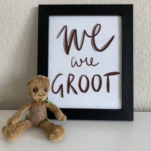 May include: A black framed picture with the text "We are Groot" in brown lettering. A brown plush toy of the character Groot from the Guardians of the Galaxy movie series is in the foreground.