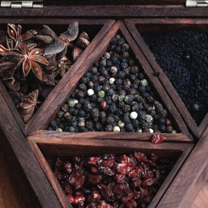 May include: A wooden box with six compartments, each containing a different spice. The compartments are arranged in a triangular pattern. The spices include star anise, black peppercorns, white peppercorns, green peppercorns, black sesame seeds, and dried cranberries.