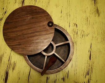 Op de afbeelding: Een ronde houten kruidenbox met een deksel en een kleine houten lepel. De box is verdeeld in secties en heeft een donkerbruine afwerking. Het deksel is gedeeltelijk open en onthult de binnenkant. De achtergrond is een verweerd geel.