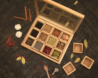 Op de afbeelding: Een houten specerijenbox met een doorzichtig deksel, open om compartimenten te laten zien die gevuld zijn met verschillende specerijen. De doos staat op een donkerbruin oppervlak met kaneelstokjes, teentjes knoflook en rode pepers die eromheen verspreid liggen.