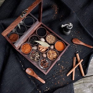 May include: A wooden box with a glass lid filled with various spices, including black peppercorns, cloves, cinnamon sticks, and bay leaves. The box is open and the spices are visible. There are also two wooden spoons and a small metal grinder on a black fabric background.