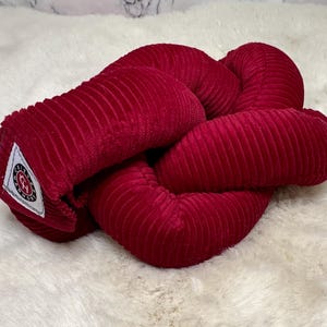 May include: A red corduroy knot pillow with a white label that says "Cozy Knots".