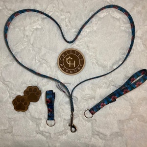 May include: A blue and red patterned dog leash with a matching shorter leash and two wooden paw prints. The leash is in the shape of a heart and has a metal clip. The wooden paw prints have the text "Critter Harmony" on them.