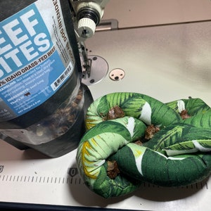 May include: A close-up of a bag of 100% Idaho grass-fed beef bites for dogs. The bag is open and the beef bites are visible. The bag is sitting on a white surface next to a green and white knotted fabric toy.