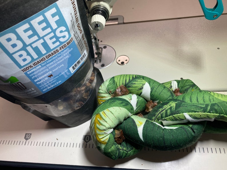 May include: A close-up of a bag of 100% Idaho grass-fed beef bites for dogs. The bag is open and the beef bites are visible. The bag is sitting on a white surface next to a green and white knotted fabric toy.