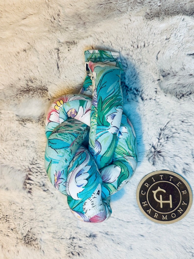 May include: A knotted, teal blue and white floral fabric dog toy with a brown wooden tag that says "Critter Harmony" in a stylized font.