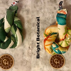 May include: Two knotted dog toys with a tropical leaf print. The toy on the left has a green and white leaf print and is labeled "Green Botanical". The toy on the right has a bright orange, yellow, and blue leaf print and is labeled "Bright Botanical". Both toys have a brown circular tag with the text "Critter Harmony" and a stylized "CH" logo.